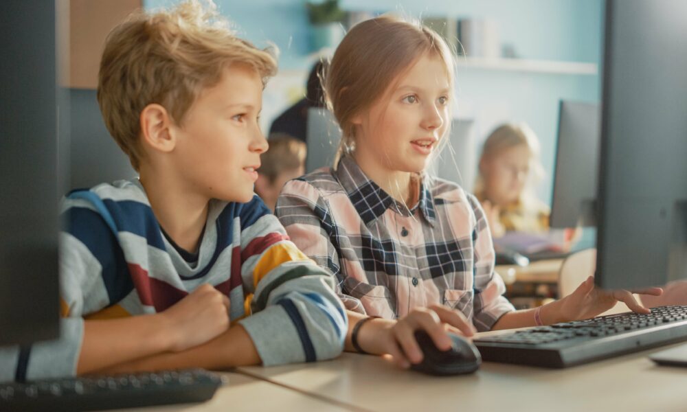 Schülerin und Schüler sitzen vor einem Computerbildschirm und bearbeiten ein interaktives Arbeitsheft